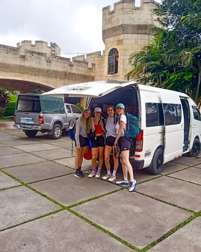 Viajeros posando junto a una van de shuttle abierta frente a un edificio estilo castillo